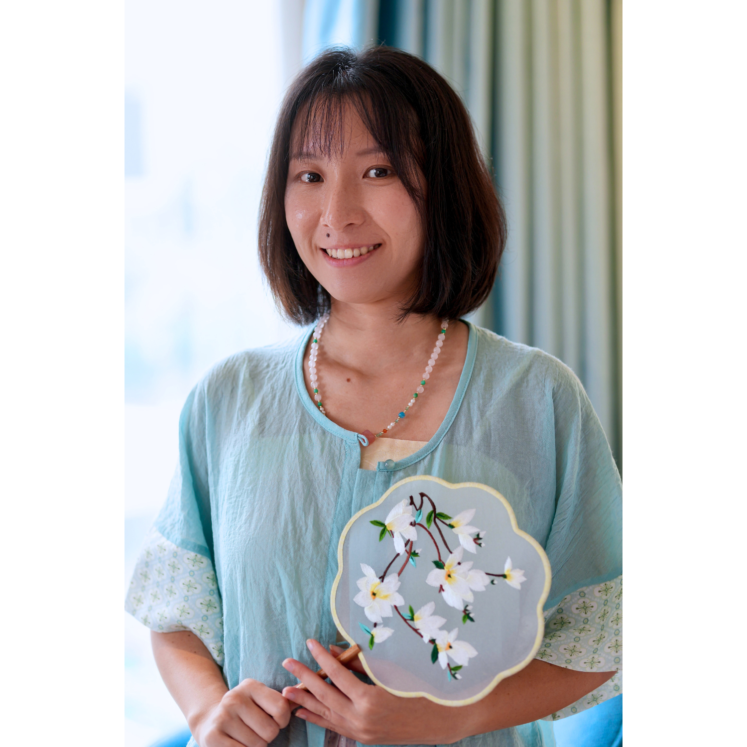 Woman holding a embroidered fan with floral design in a light blue shirt.